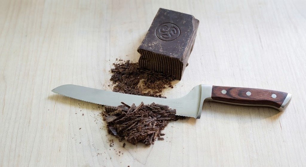 A block of dark chocolate sits beside chocolate shavings and a serrated knife on a light wooden surface.