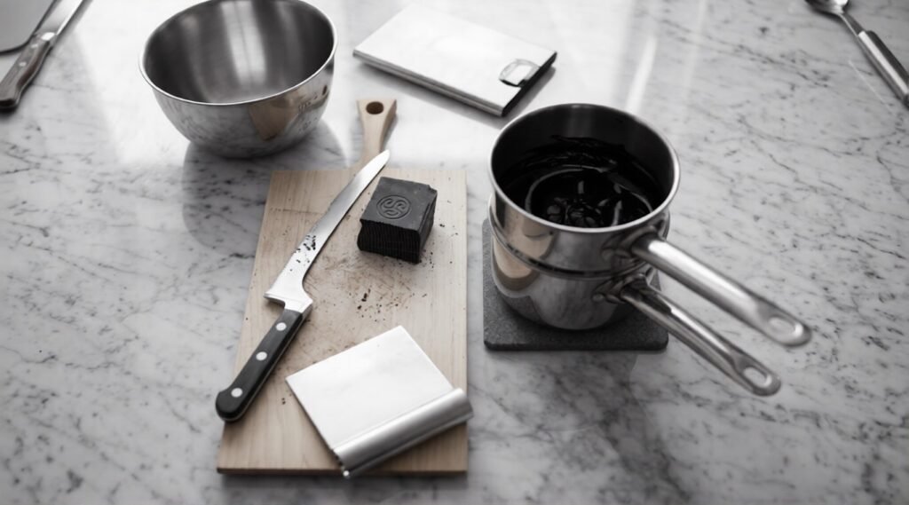Chocolate-melting tools are arranged on a marble counter, including a double boiler, stainless steel bowl, bench scraper, knife, cutting board, and block of dark chocolate.