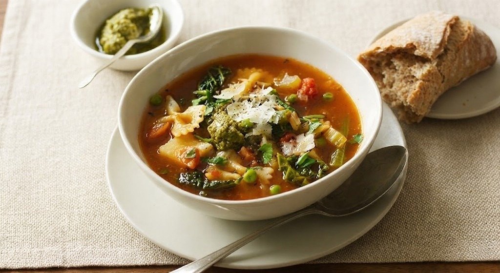 Bowl of Mediterranean lentil and vegetable soup with pasta, broccoli, peas, and grated cheese, served with a spoon, a piece of crusty bread, and a small dish of green pesto.