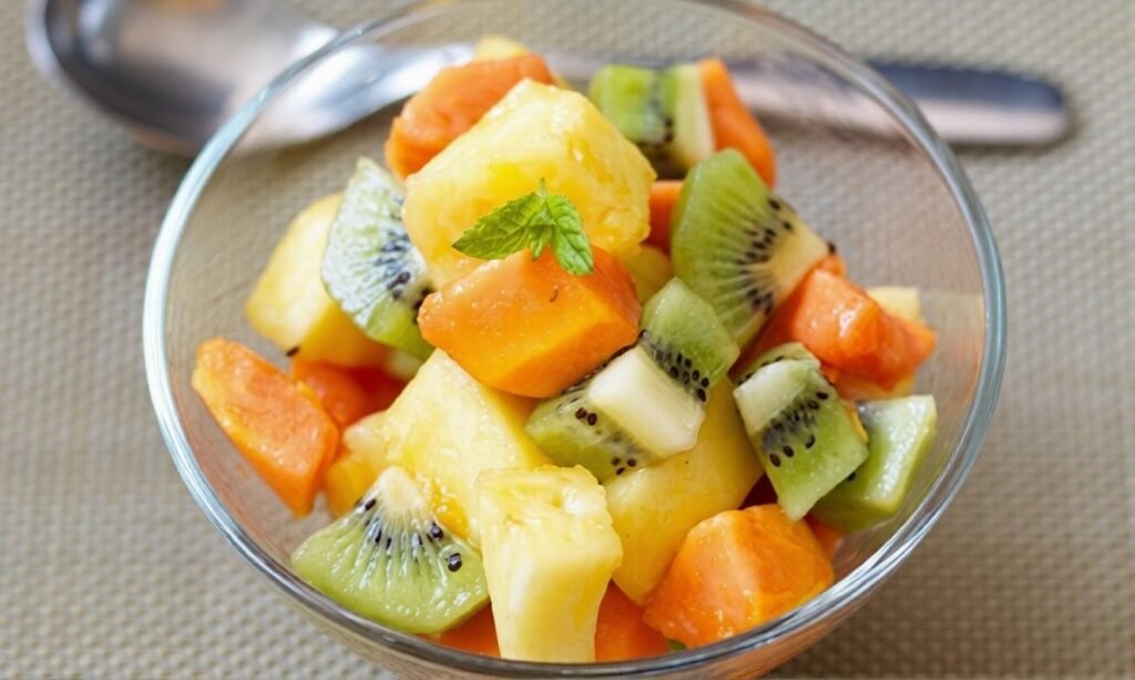 Glass bowl of mixed fruit with pineapple, kiwi, and orange pieces, topped with a small mint leaf, photographed as a light Mediterranean-style pudding or snack.
