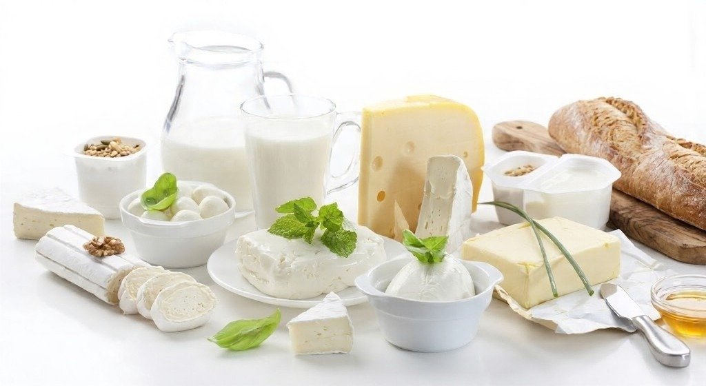 Assortment of yogurt, milk, and cheeses on a white surface, including soft and hard cheeses, butter, bread, and fresh herbs.