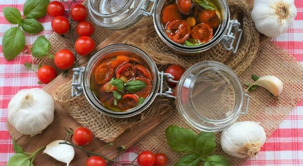 Open glass jars of preserved tomatoes with basil sit on a rustic table beside fresh cherry tomatoes, garlic, and basil leaves.