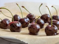 Make Homemade Chocolate Covered Cherries A dozen dark cherries with stems are half-dipped in glossy chocolate on parchment-lined plate.