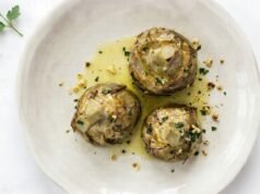 Make Roman Artichokes in an Easy Way Three cooked Roman-style artichokes sit in olive oil on a pale plate, sprinkled with herbs and bits of garlic.